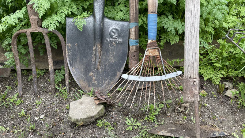 Garden spade, fork, rake and hoe leaning against a wooden fence