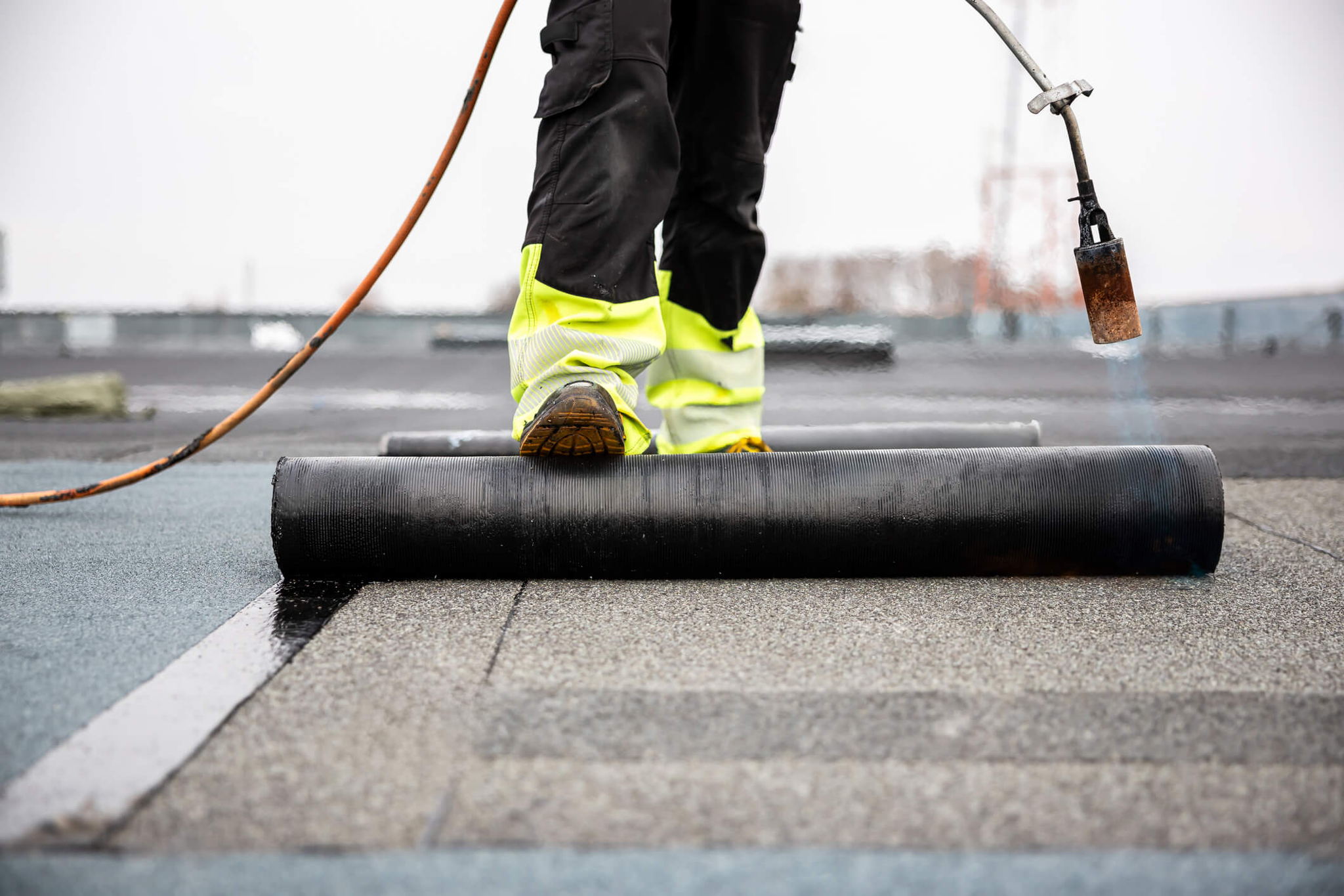 A person applying a roll of waterproof membrane to a flat roof. They are wearing bright yellow pants, and their foot is pressing down on the roll. A propane torch is used to bond the material to the surface. Rooftop and sky are visible.