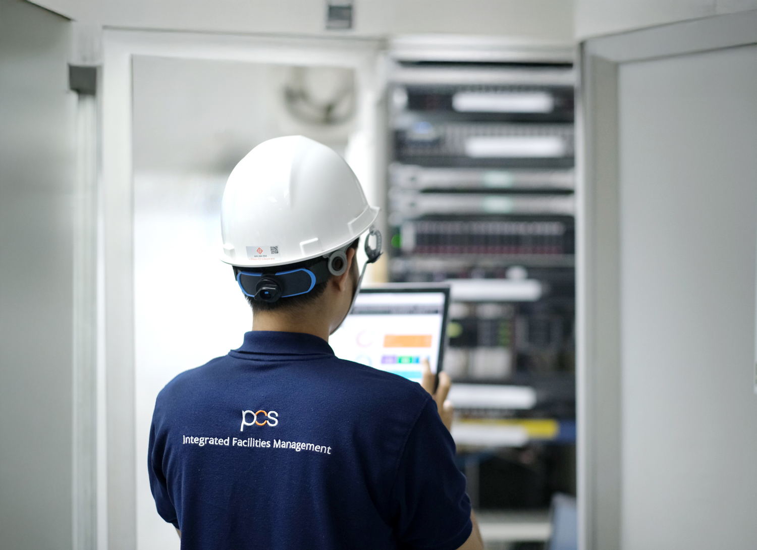 A person wearing a white hard hat and a blue Integrated Facilities Management shirt uses a tablet while standing in front of server racks in a brightly lit room.