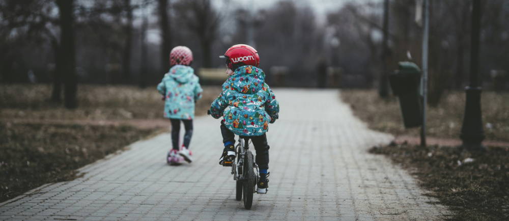 two kids out riding, one on a scooter and one on a bike wearing a woom logo on a helmet