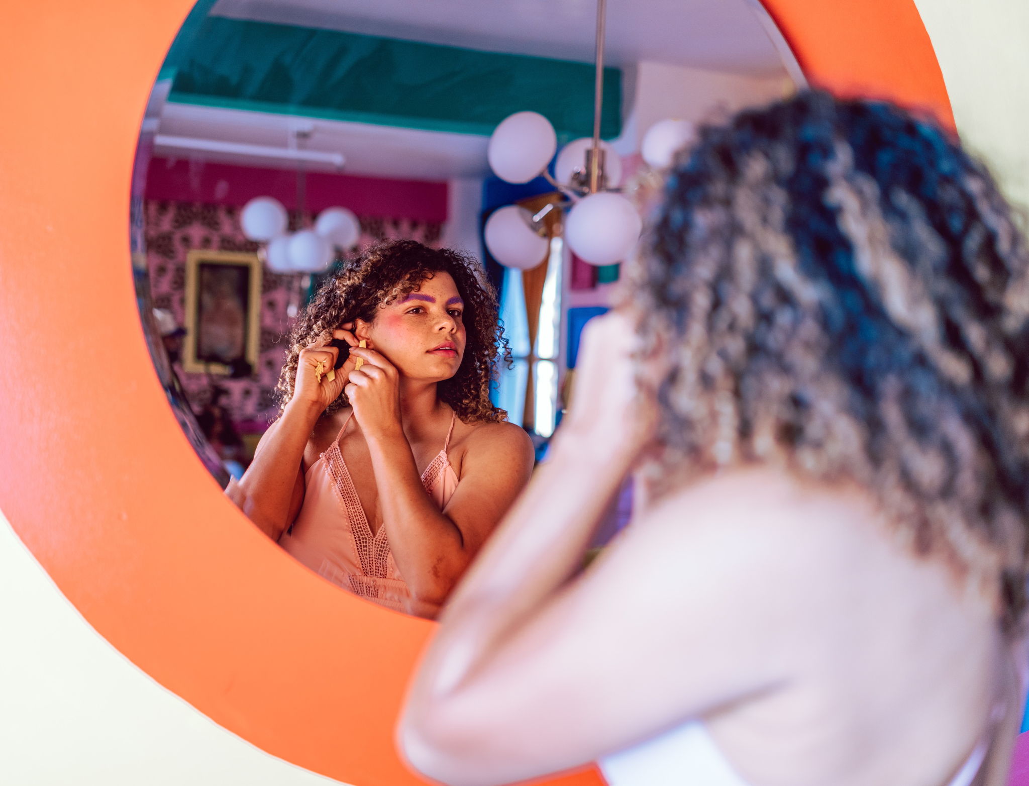 A beautiful mixed woman with curly hair looks at herself in the mirror while putting on an earring.