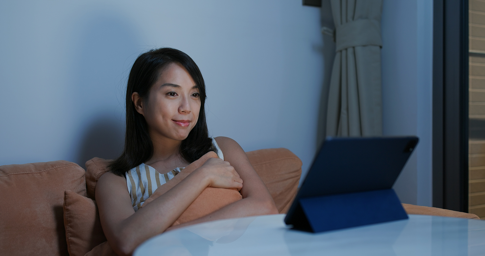 An asian woman smiles as she watches something on her device while holding a pillow.