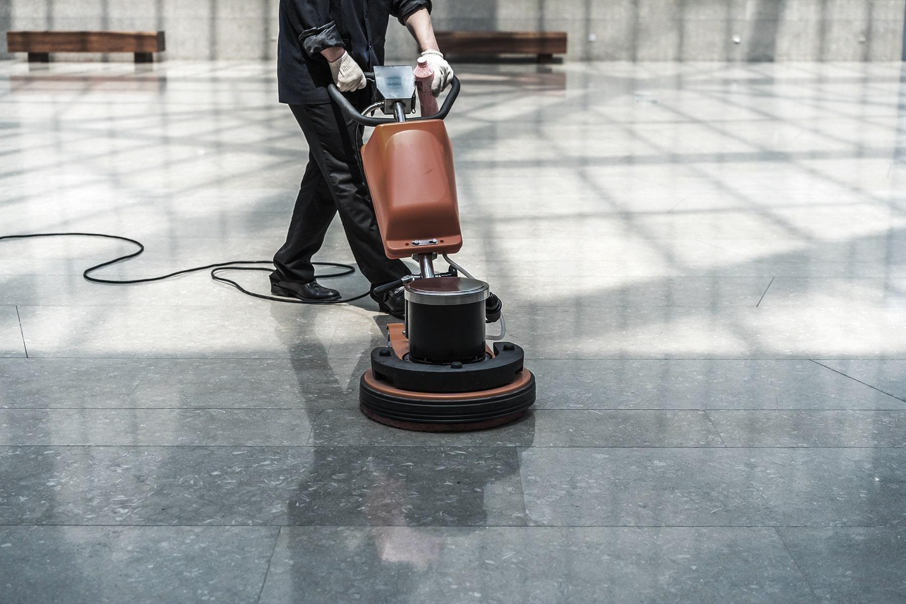 A person operating an industrial floor cleaning machine on a shiny, reflective, gray marble floor inside a large, well-lit building. Only the persons lower body and hands are visible.