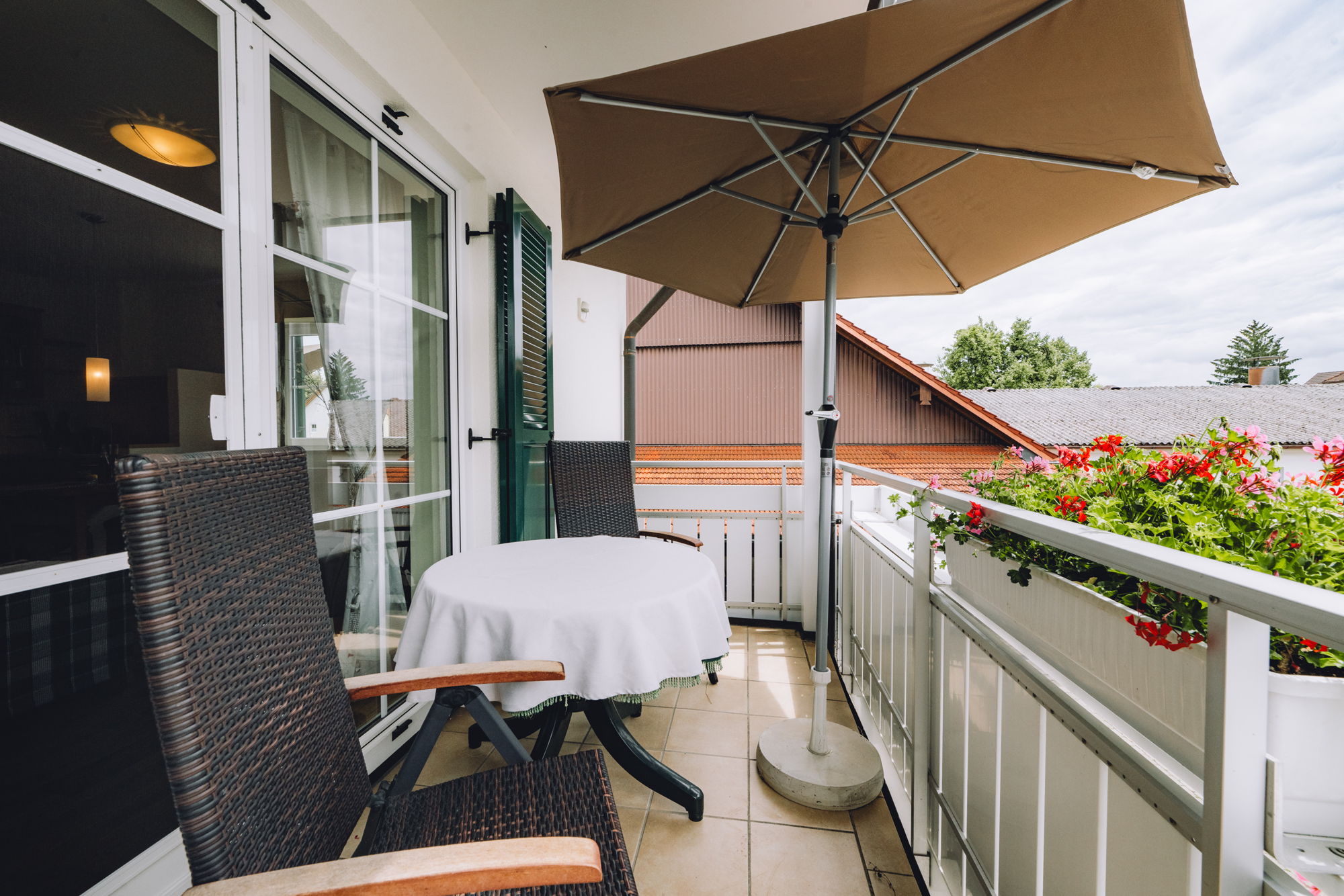 Ein Balkon mit Sonnenschirm, weißem Tisch, verstellbaren Stühlen und Blumenkasten.