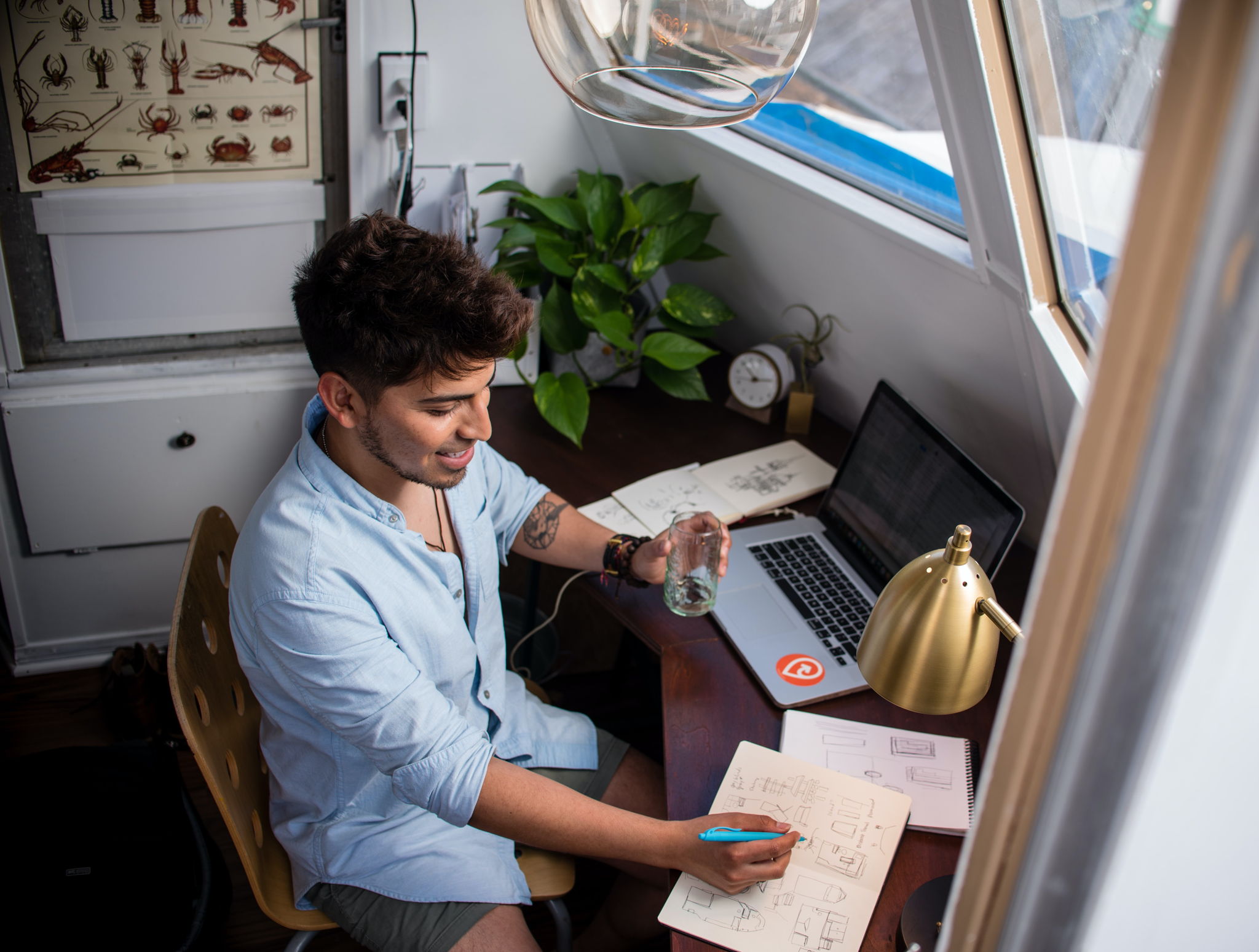 A young hip guy sits on his desk with his laptop and writes something on a note pad. He is smiling and holding a glass of water.