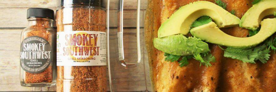 Two bottles of FreshJax Organic Smokey Southwest Grill Seasoning next to a dish filled with enchiladas topped with avocados and cilantro. 