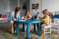 Kids sitting at a table and drawing while supervised by a teacher. 