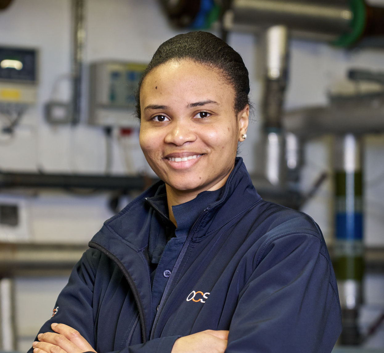 A woman wearing a navy blue jacket stands indoors with her arms crossed, smiling. The background shows industrial pipes and equipment in a workplace setting.