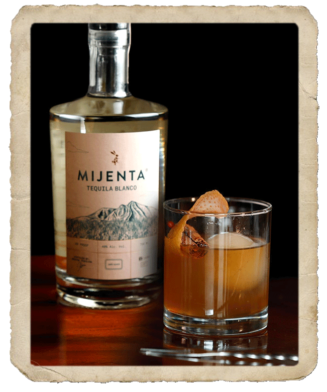 Tequila cocktail with orange peel and cinammon stick in the foreground and Mijenta Tequila Blanco bottle in the background