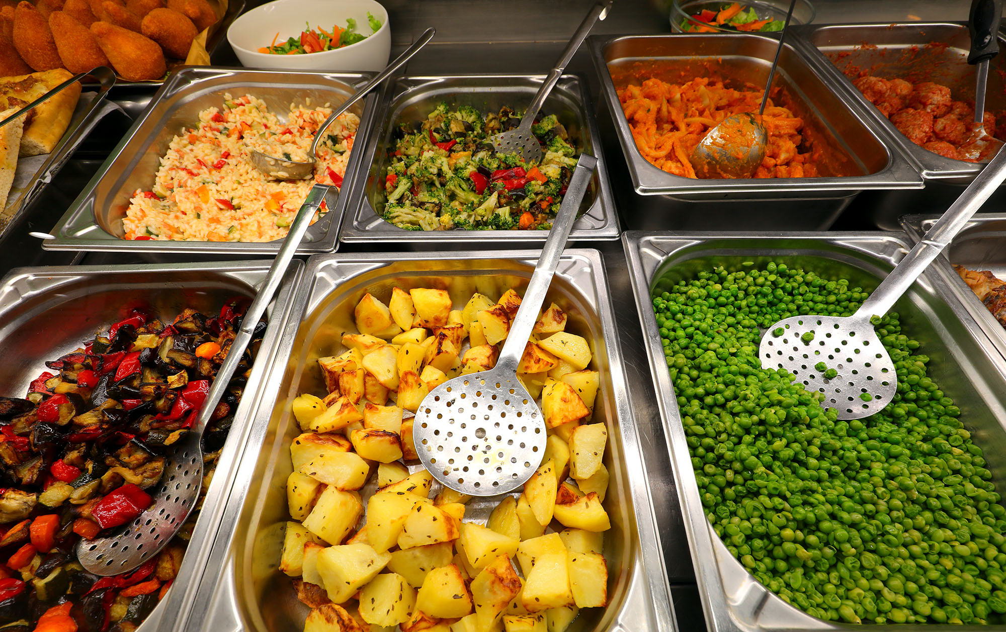 A buffet with trays of assorted hot foods, including roasted potatoes, green peas, rice, mixed vegetables, and other colorful dishes, each with a metal serving spoon or slotted spoon.