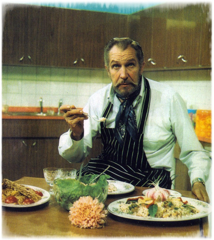Vincent Price from on of the pages of his cookbook, he has an apron and a spoon in hand, with several plates full of food in front of him.