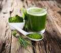 a short glass of wheat grass juice surrounded by fresh wheat grass and dark green wheat grass powder, on a rustic wood background