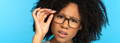 Model wearing glasses infront of a light blue background, holding the arm of her glasses, looking confused