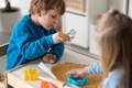 Two children use colorful cookie cutters to shape dough on parchment paper, engaging in a fun and creative baking activity at a play table.