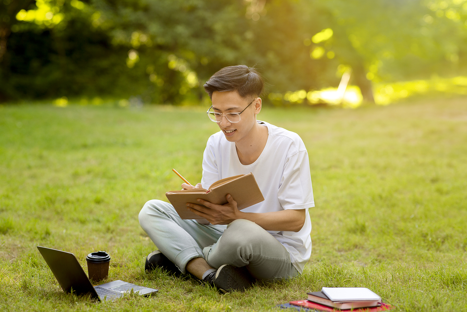 Focused Asian Student Guy Preparing For Lessons Outdoors, Reading Book And Taking Notes While Sitting On Grass In Park