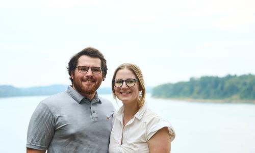 Two smiling individuals stand by a serene body of water, conveying warmth and joy in a beautiful natural setting.