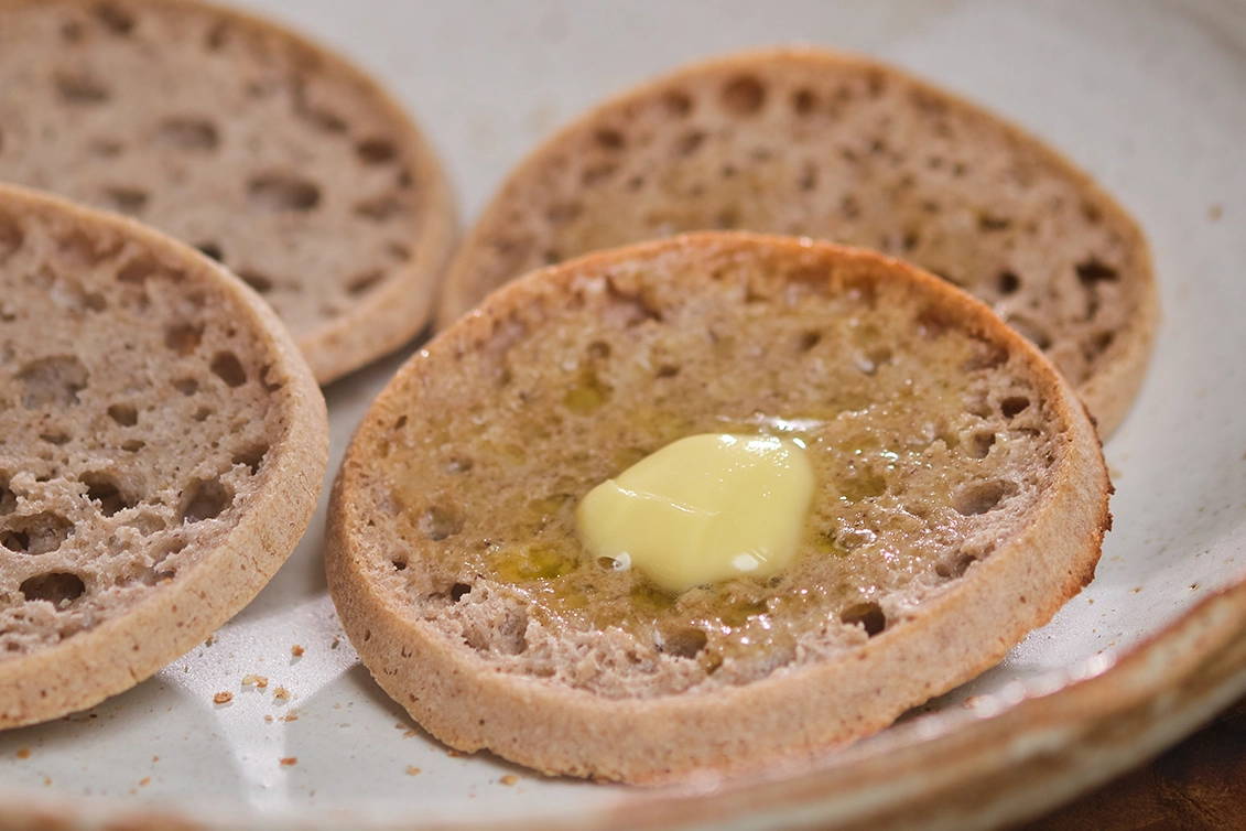 Four toasted English muffin halves arranged on a ceramic plate, with a dab of melting butter on the muffin in the foreground.