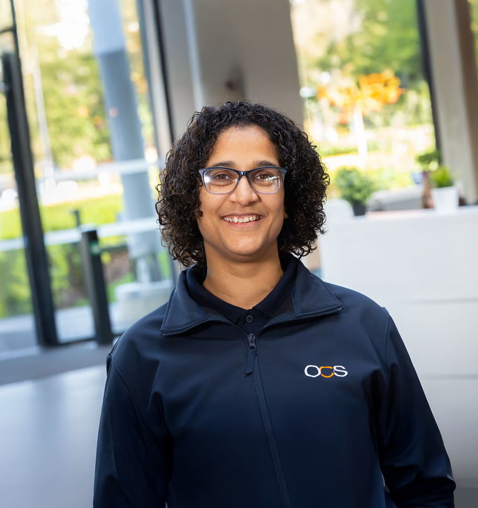 A person with short curly hair and glasses, smiling and wearing a dark jacket with an OCS logo, stands indoors in a bright, modern space with large windows and greenery outside.