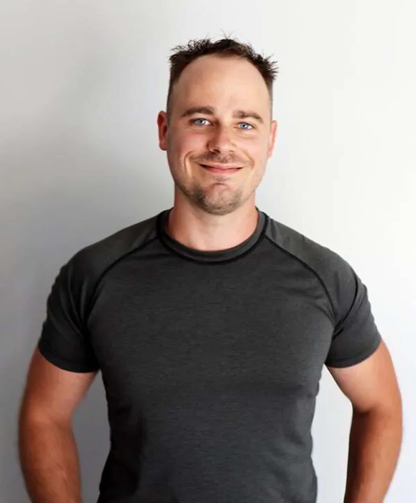 Smiling man with short hair wearing a dark crew-neck athletic shirt, standing against a light background.