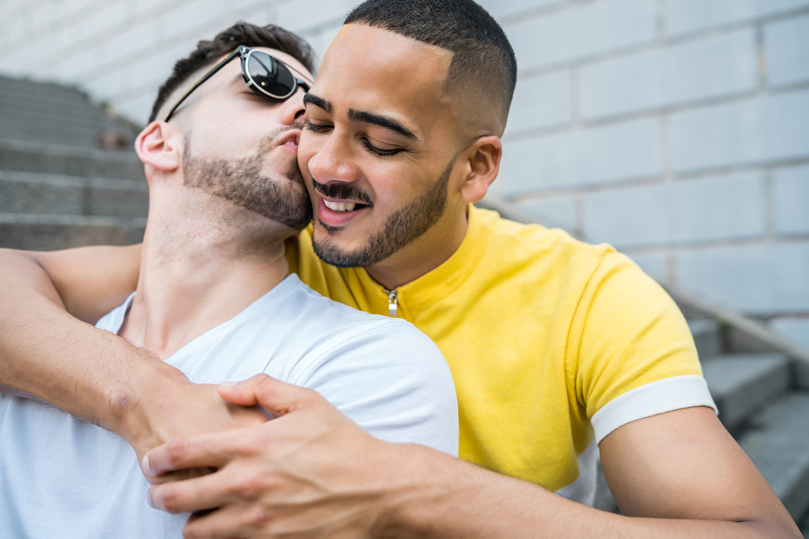 Two multi ethnic young men kiss. One has his arms wrapped around the other smiling.