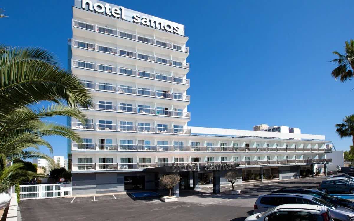 Modernes mehrstöckiges Hotelgebäude mit Balkonen, Palmen und Parkplatz bei strahlend blauem Himmel.