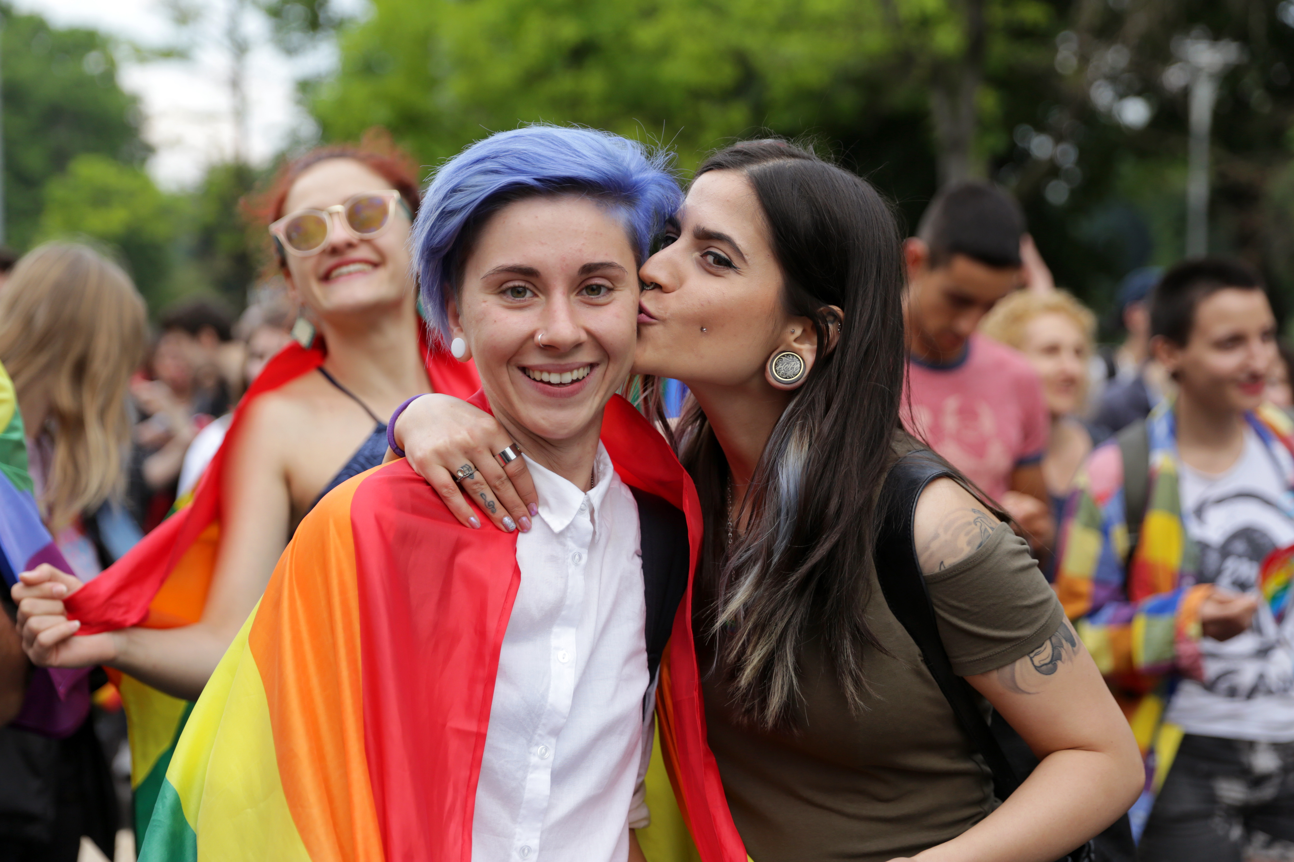 Two young and hip women with stylish outfits and hair smile for the camera and while one kisses the others' cheek. Her friend is wearing a pride flag.