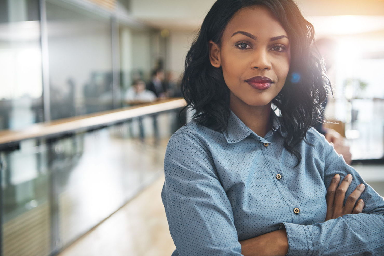 A confident black woman crosses her arms while smiling looking at the camera. She is in an office with people working behind her.