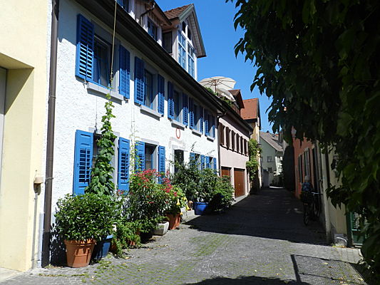  Konstanz
- Blick auf ein gemütliches Gässchen in der Konstanzer Altstadt. Eine Hausfassade sticht heraus, da sie weiß ist und blaue Fensterläden hat. Außerdem ist es reich mit Blumen geschmückt.