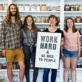 Beth and three other people stand in front of spice shelves while holding a sign that says 'Work Hard and Be Nice to People'.