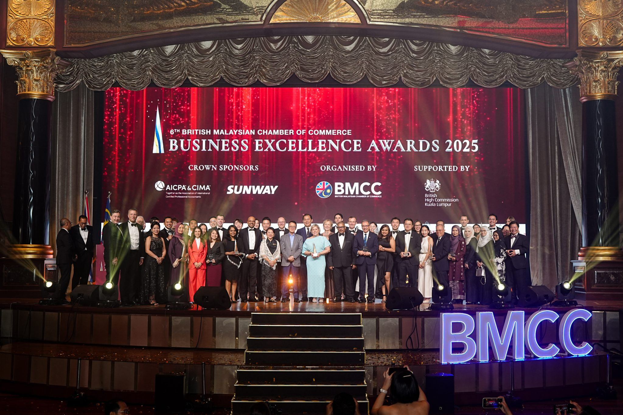 A large group of people in formal attire stand on a decorated stage at the British Malaysian Chamber of Commerce Business Excellence Awards 2025, with banners and logos displayed in the background.