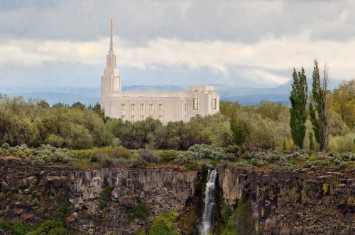 Twin FAlls TEmple standing on a hilltop with a waterfall tumbling dowin the clifff ace. 