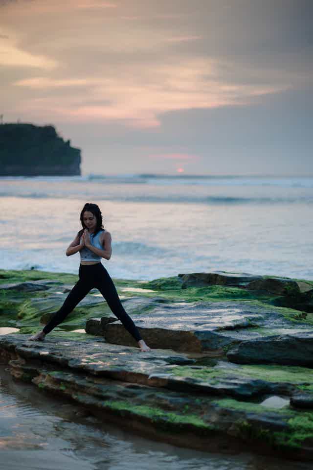 Woman practicing yoga on rocky shoreline at sunset, balancing in Warrior Pose with mossy stones and calm sea.