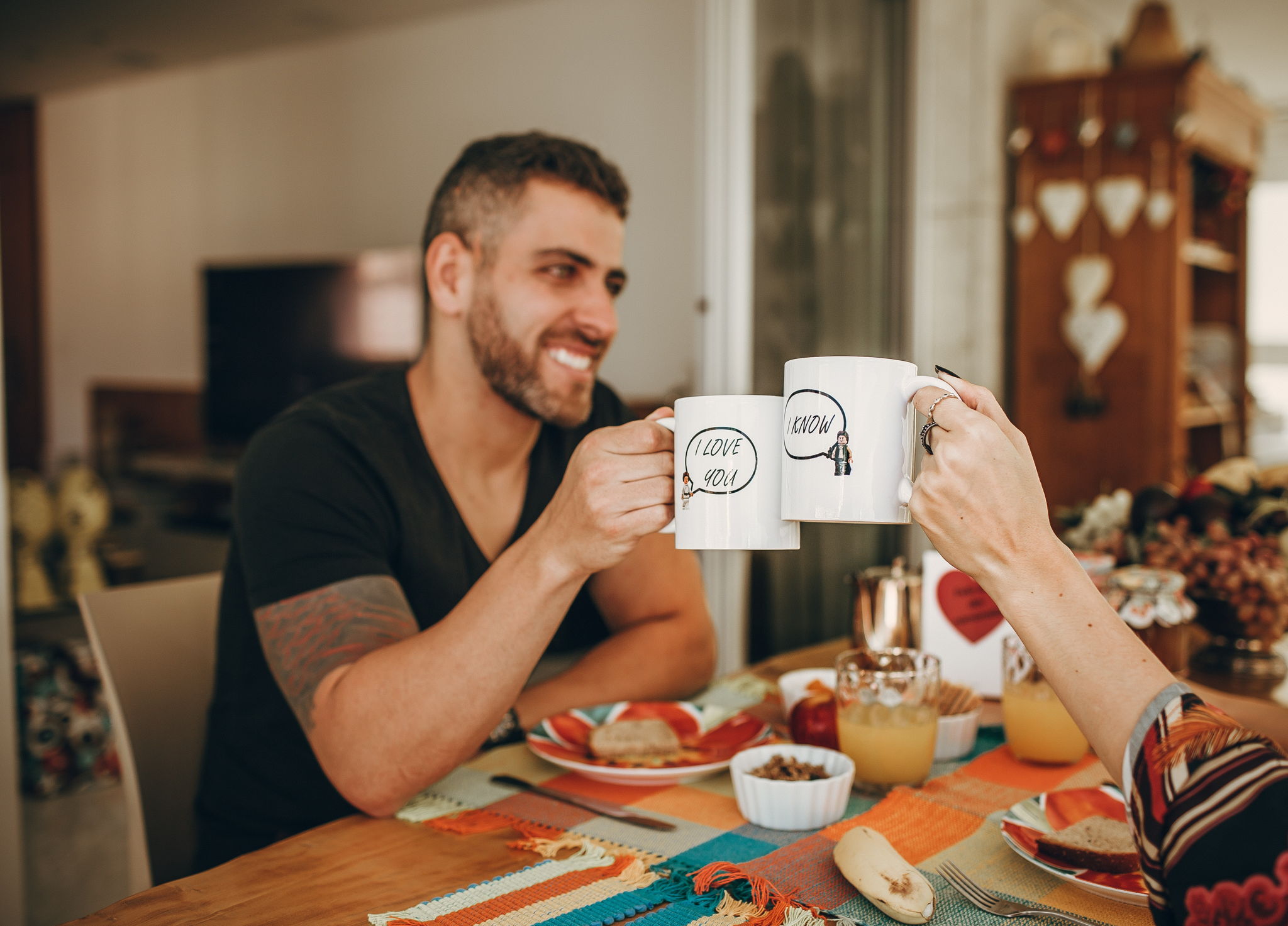 Image of an attractive man sitting with a woman out of frame having breakfast together. He is smiling and holding a white coffee mug up to the woman's coffee mug.