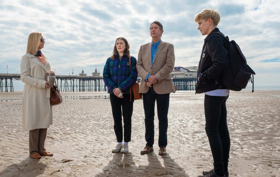 Mae speaking with her family with her backback on, on a sandy beach. Everyone looks uncomfortable.