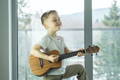An image of a young boy playing the ukuele with a a satisfied look on his face. He is in front of a large window that and there is a calm, cool day outside. 
