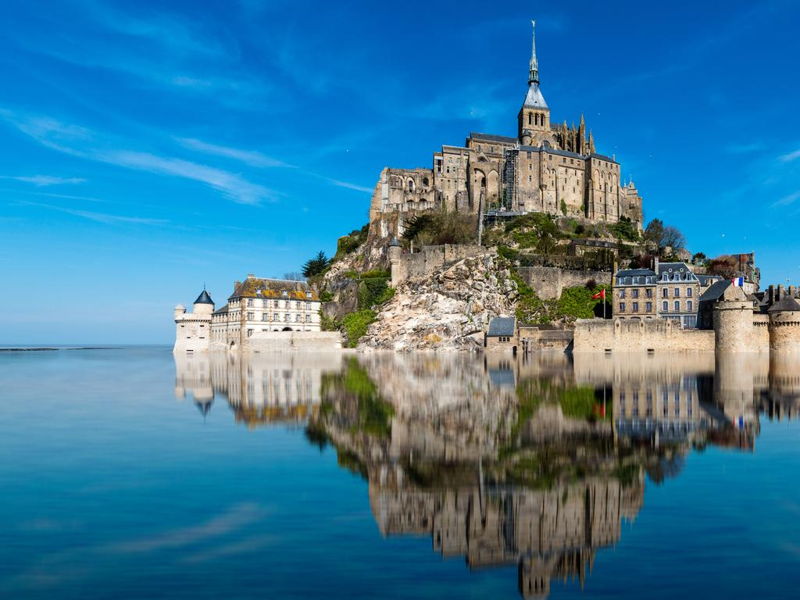 Vue majestueuse du Mont Saint-Michel se reflétant dans l'eau à marée haute, sous un ciel bleu éclatant
