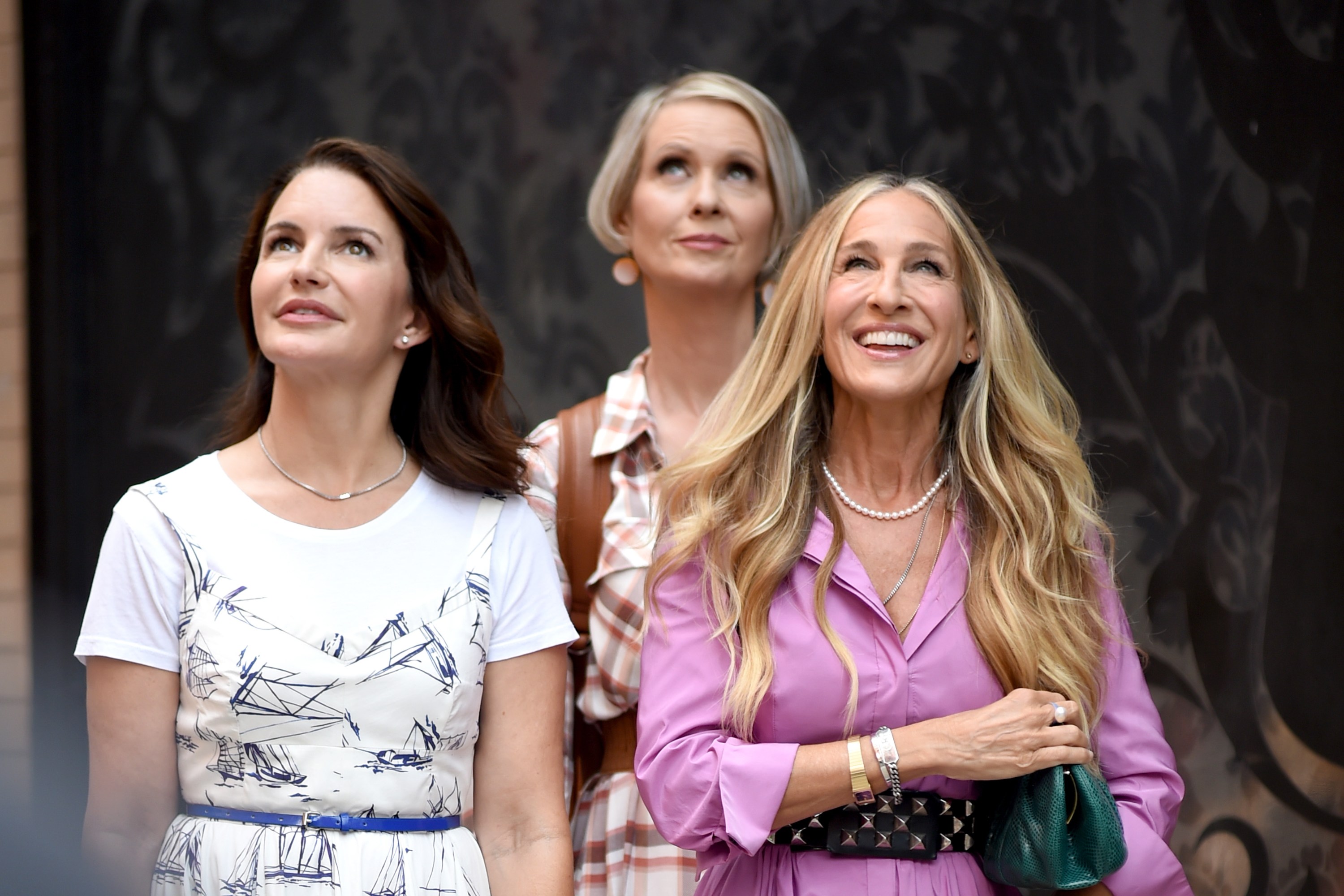 Miranda, Samantha, and Carrie walk together in the streets. They are all looking up at something out of frame and are smiling together.