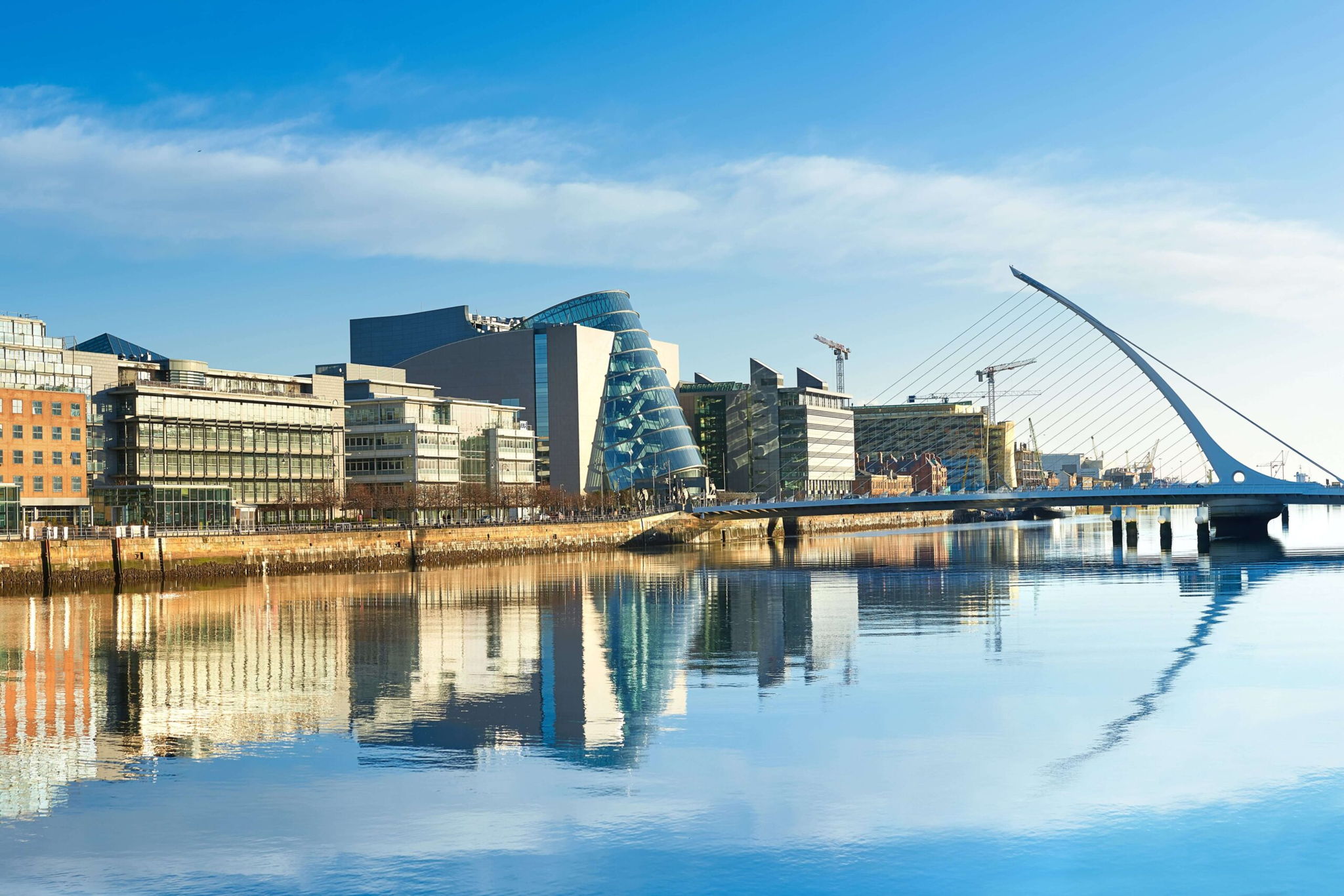 Skyline of a modern city along a clear river, featuring distinctive architecture including a building with a spiral design and a harp-shaped bridge. The buildings and bridge are mirrored in the calm water under a bright blue sky.