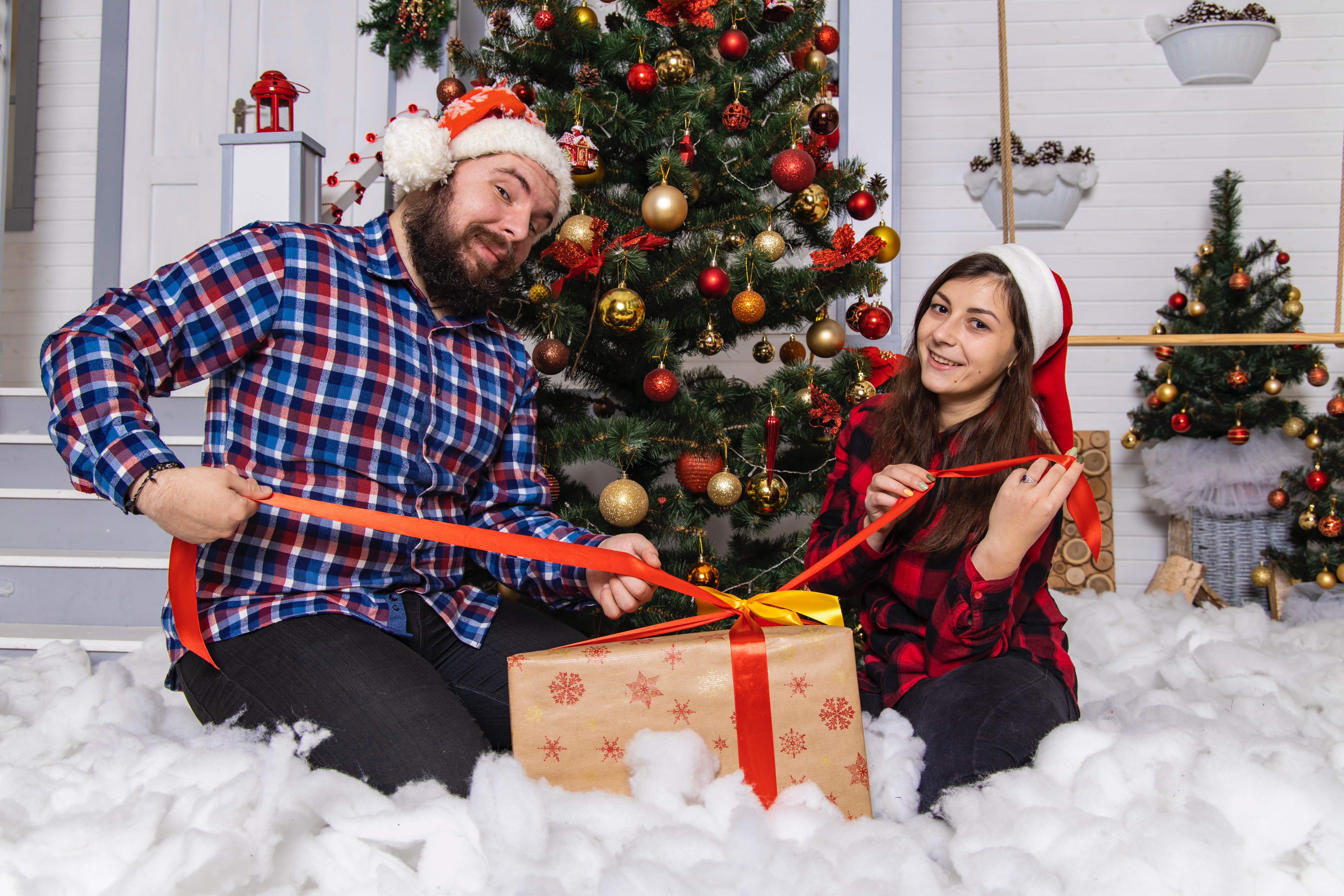 A man and woman both wearing santa hats, wrap a christmas present while infront of a tree smiling.