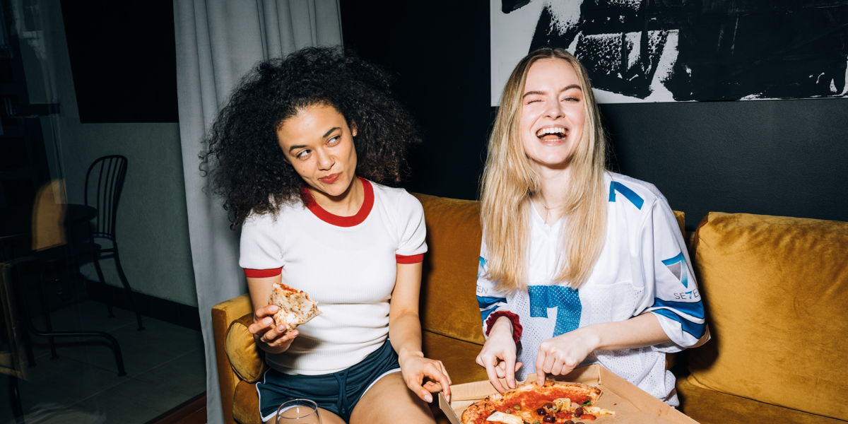Two friends, a black woman and a white woman both eating pizza and laughing with smirks.