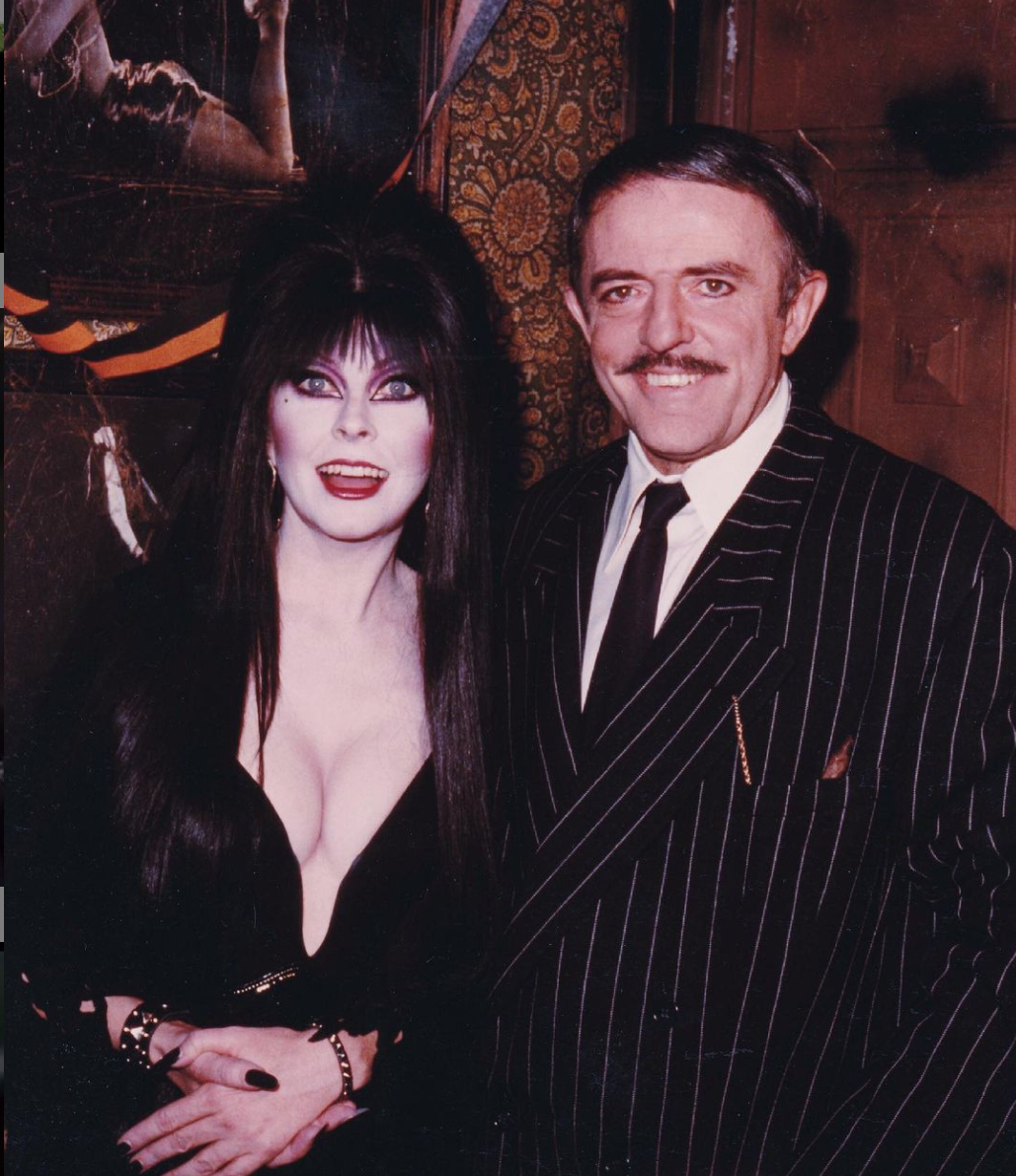 Elvira posing for a photo with Gomez Addams both in their costumes smiling.