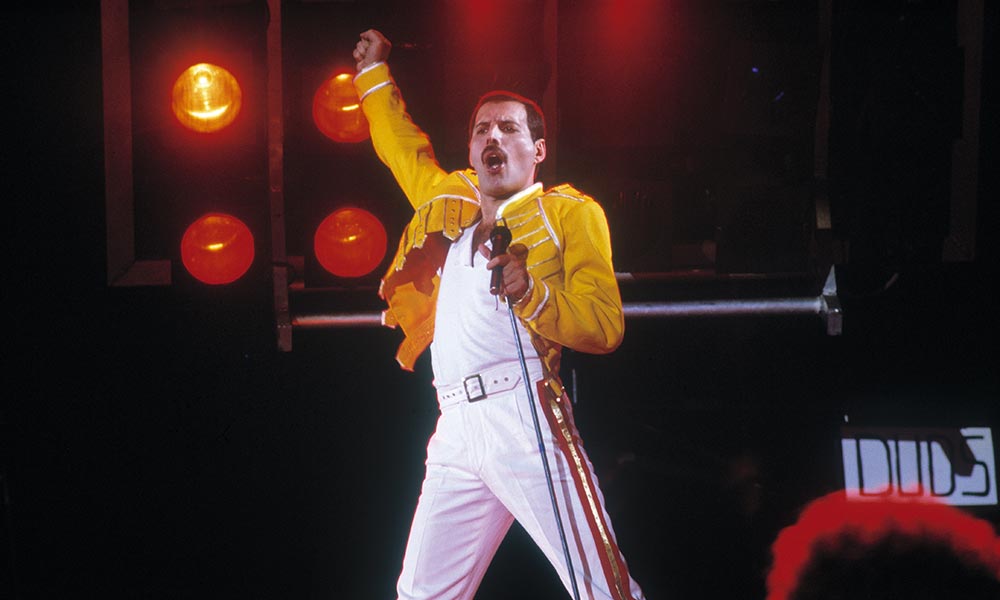 Freddie onstage holding the mic stand and his other hand in a fist in the air singing.