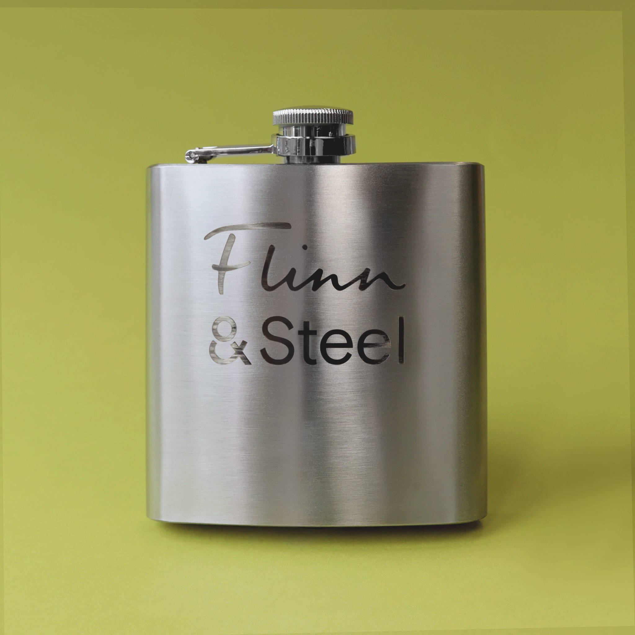 A close up image of a stainless steel hip flask on a lime green background. The flask is stood up with the top closed and a Flinn & Steel logo engraving on the side.