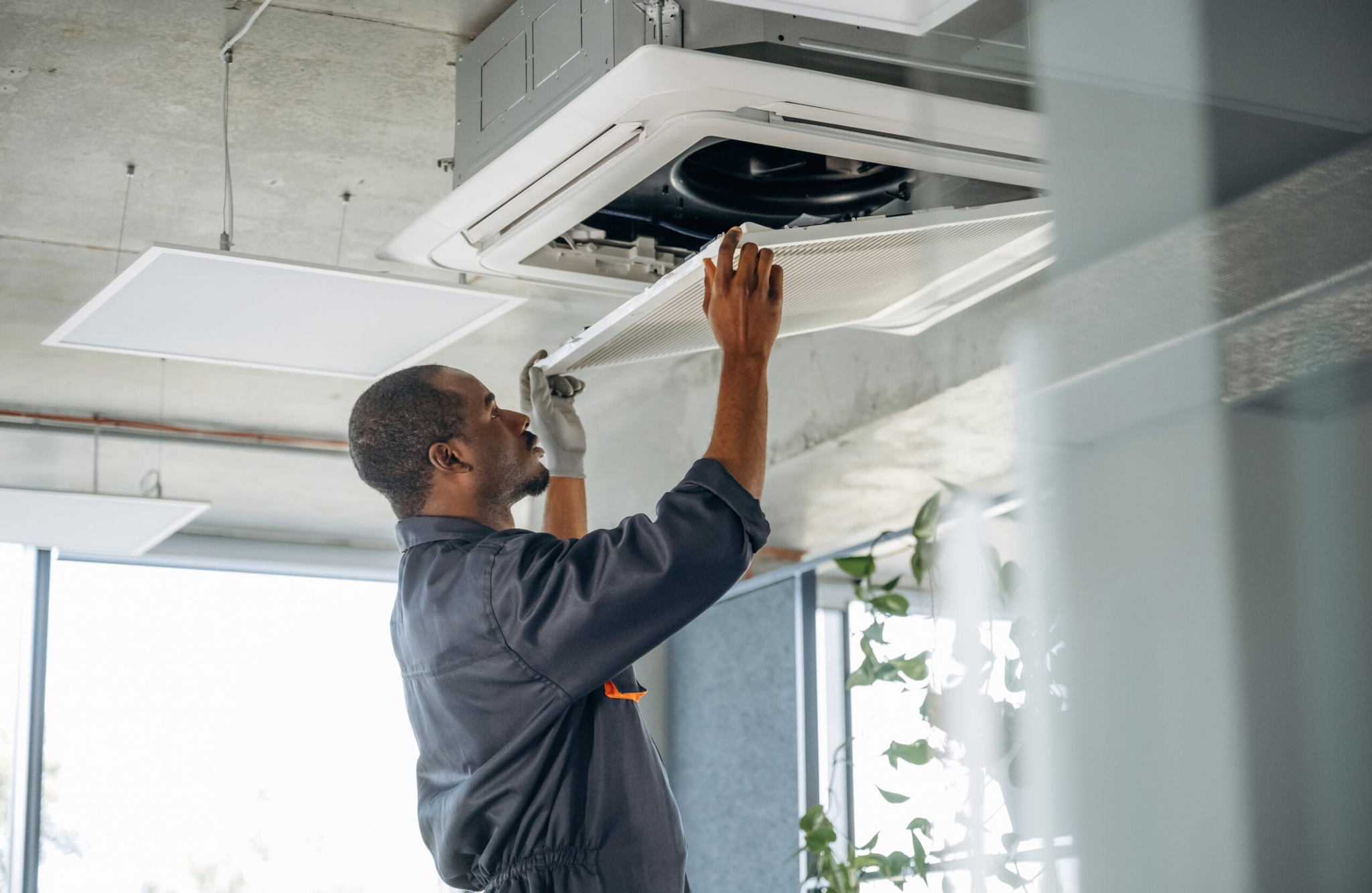 A maintenance worker in a gray uniform and gloves inspects an overhead air conditioning unit in a modern office setting. The worker is adjusting or replacing a component, with visible plants and large windows in the background.