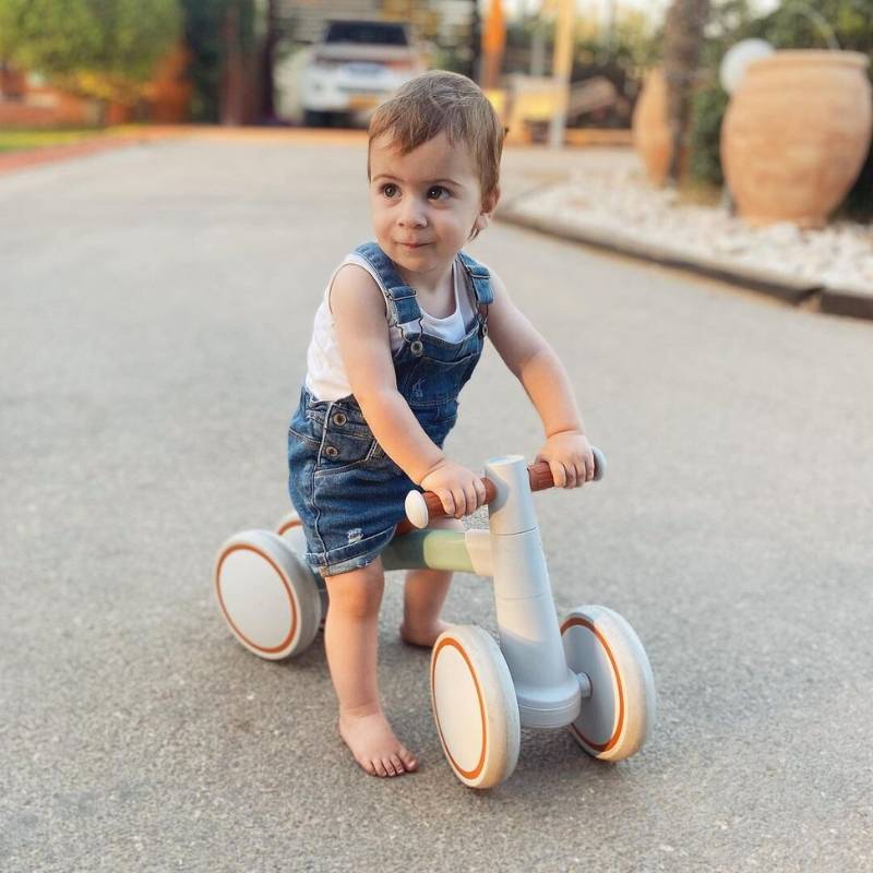 Un tout-petit en salopette en jean monte un vélo d'équilibre pour bébé InvenToy Montessori sur un chemin pavé, regardant en arrière avec un léger sourire, près d'un grand pot en céramique et de verdure en arrière-plan.