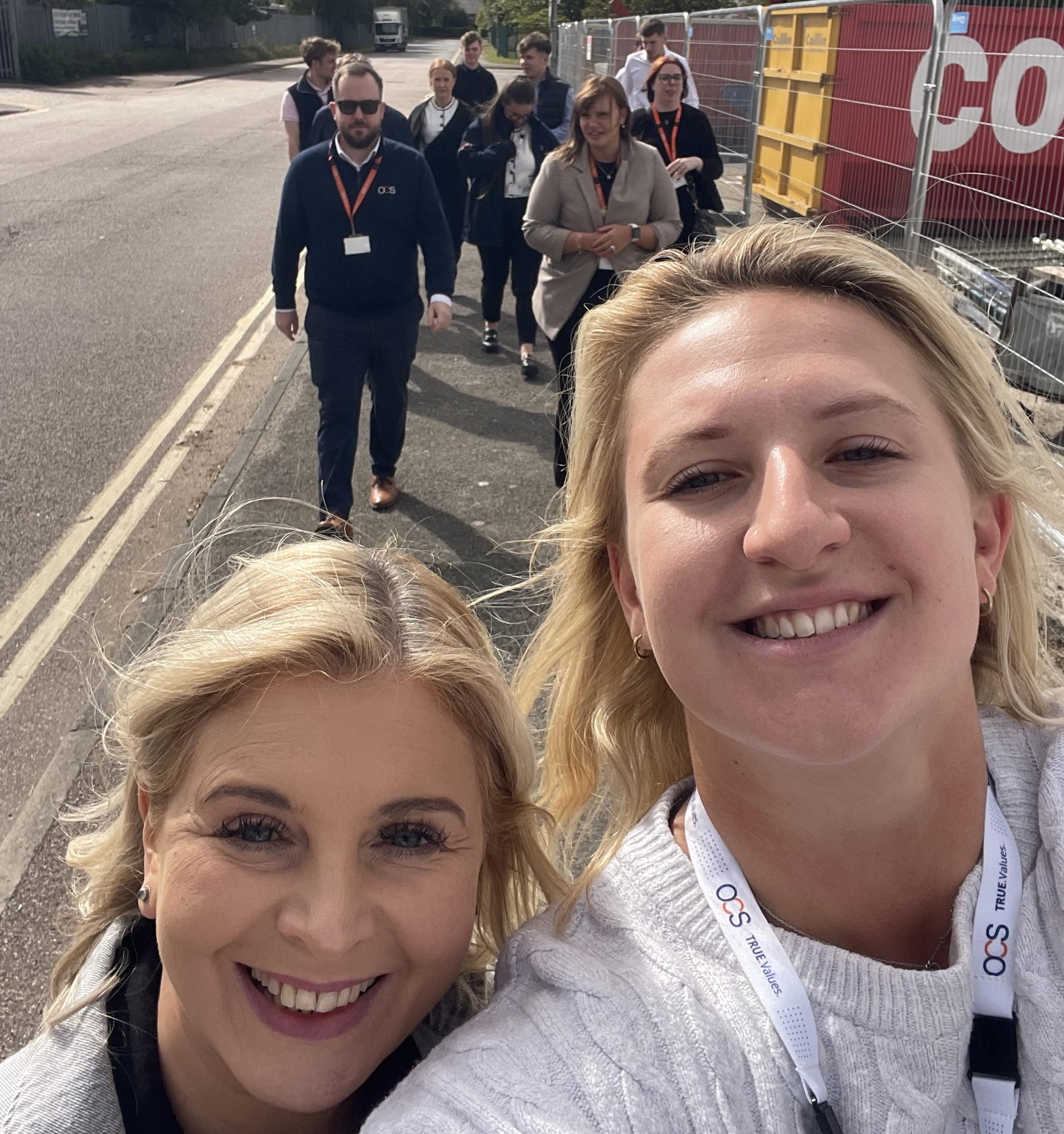 A group of people, most wearing lanyards, walk along a sidewalk on a sunny day. Two smiling women are in the foreground, taking a selfie, while others follow behind them.