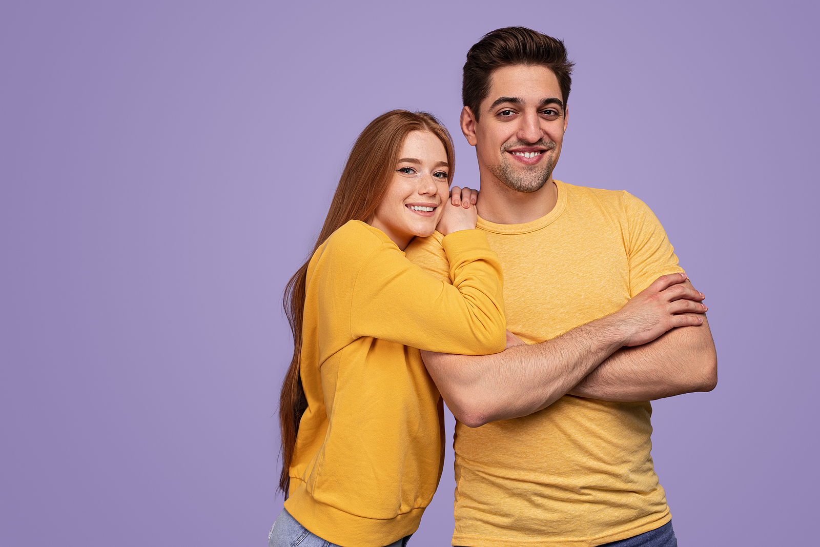 A young attractive man and woman smile and pose with arms crossed against a purple background.