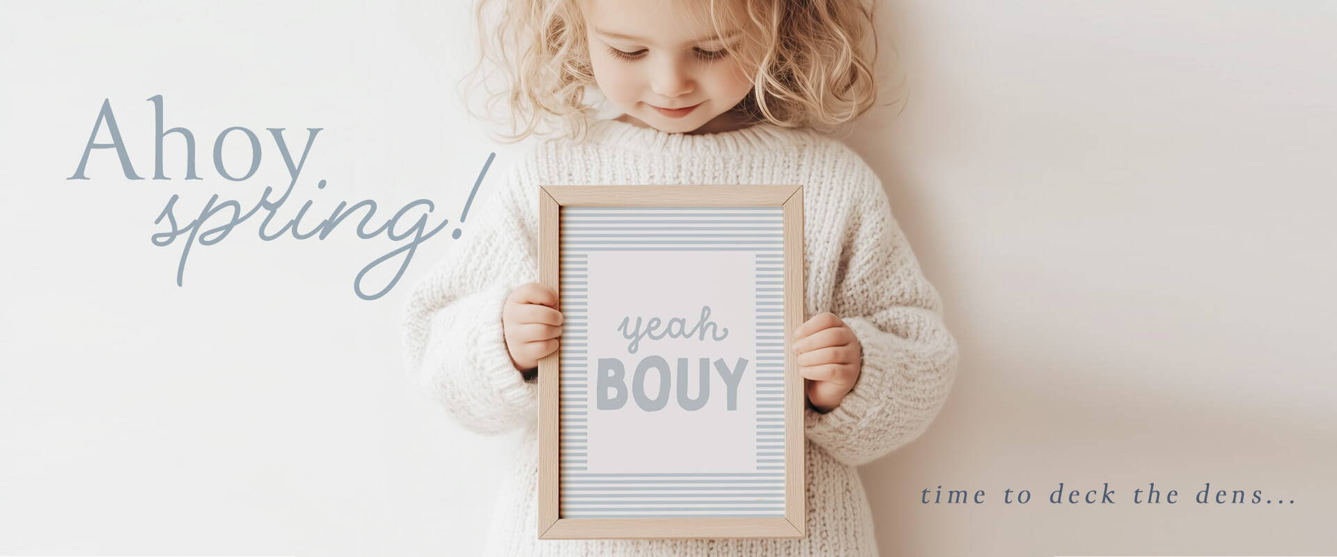 Cheerful toddler in a cozy white sweater holding a 'Yeah Bouy' framed artwork with nautical stripes, under the phrase 'Ahoy, spring! Time to deck the dens...' – perfect for springtime coastal decor.