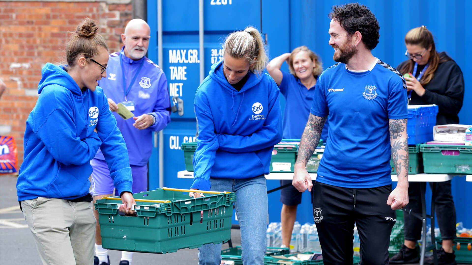 A group of volunteers from Everton in the Community wearing blue shirts are smiling and organising crates outdoors in front of a blue shipping container. One person is holding a clipboard, while others chat and handle crates.