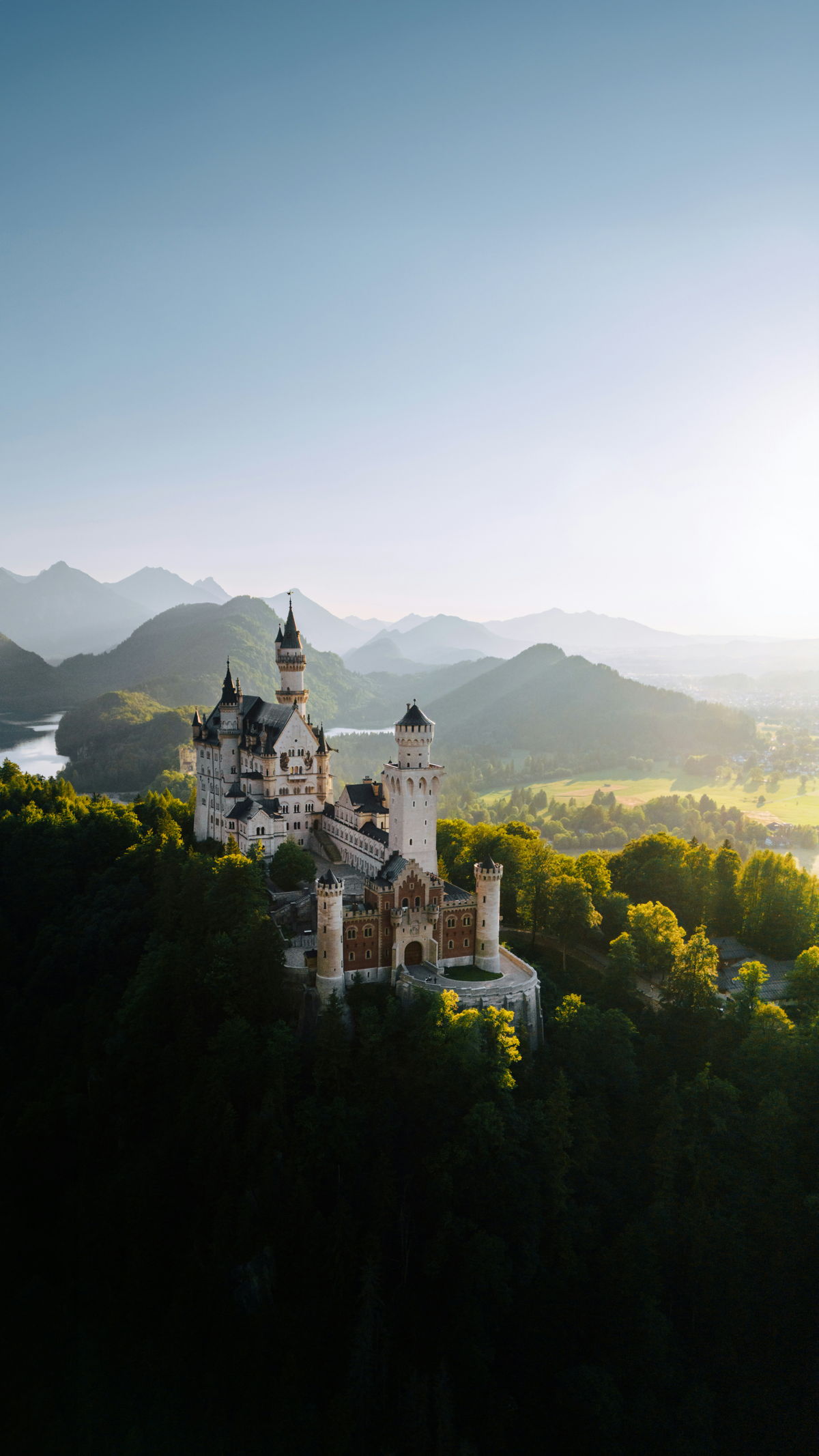 Luftaufnahme von Schloss Neuschwanstein, umgeben von grünen Wäldern, im Hintergrund Berge.